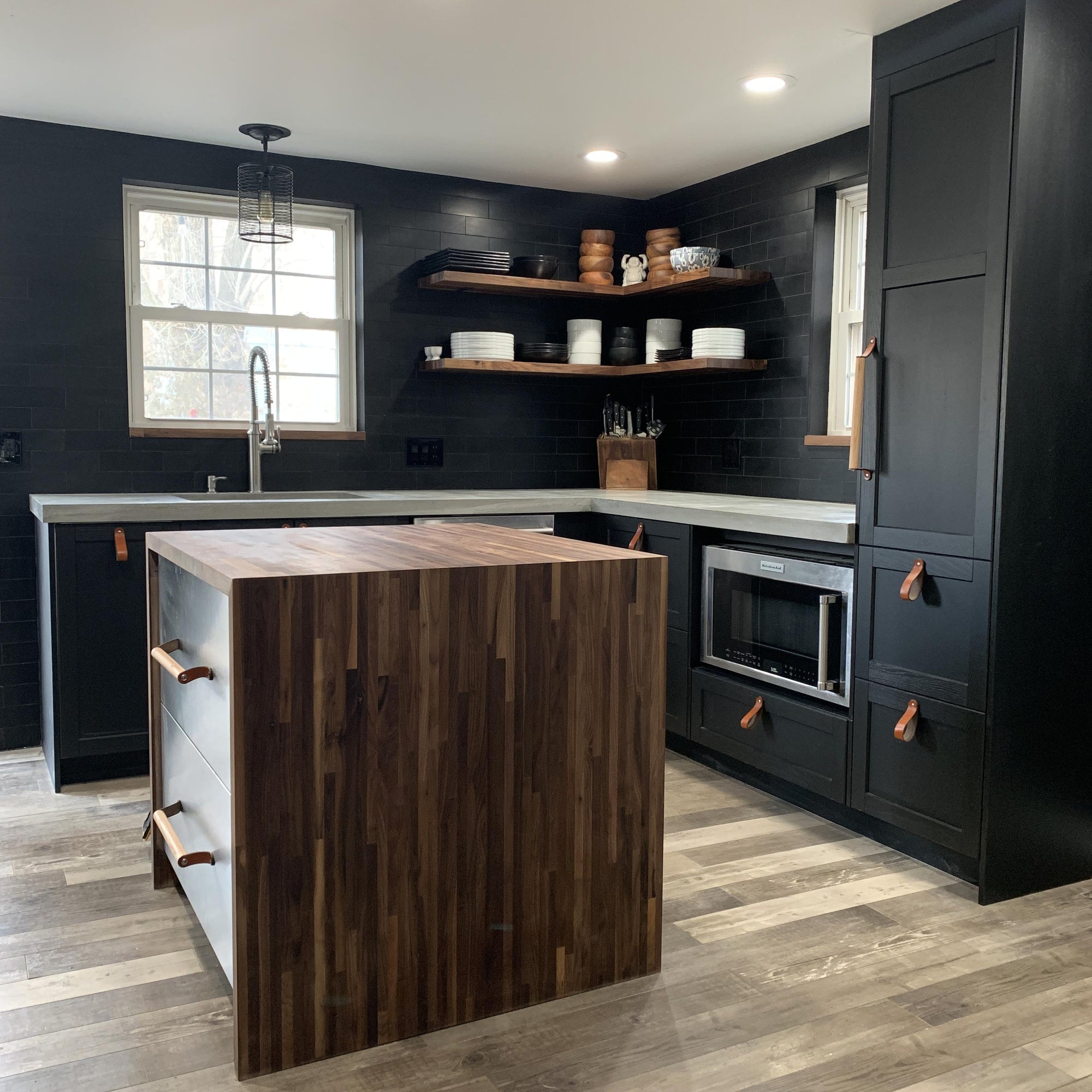 A dramatic home chef's kitchen with black-brown cabinetry, a custom spillover wood island, and stainless steel drawer fronts and accents, with honey leather and oakwood drawer pulls by Walnut Studiolo