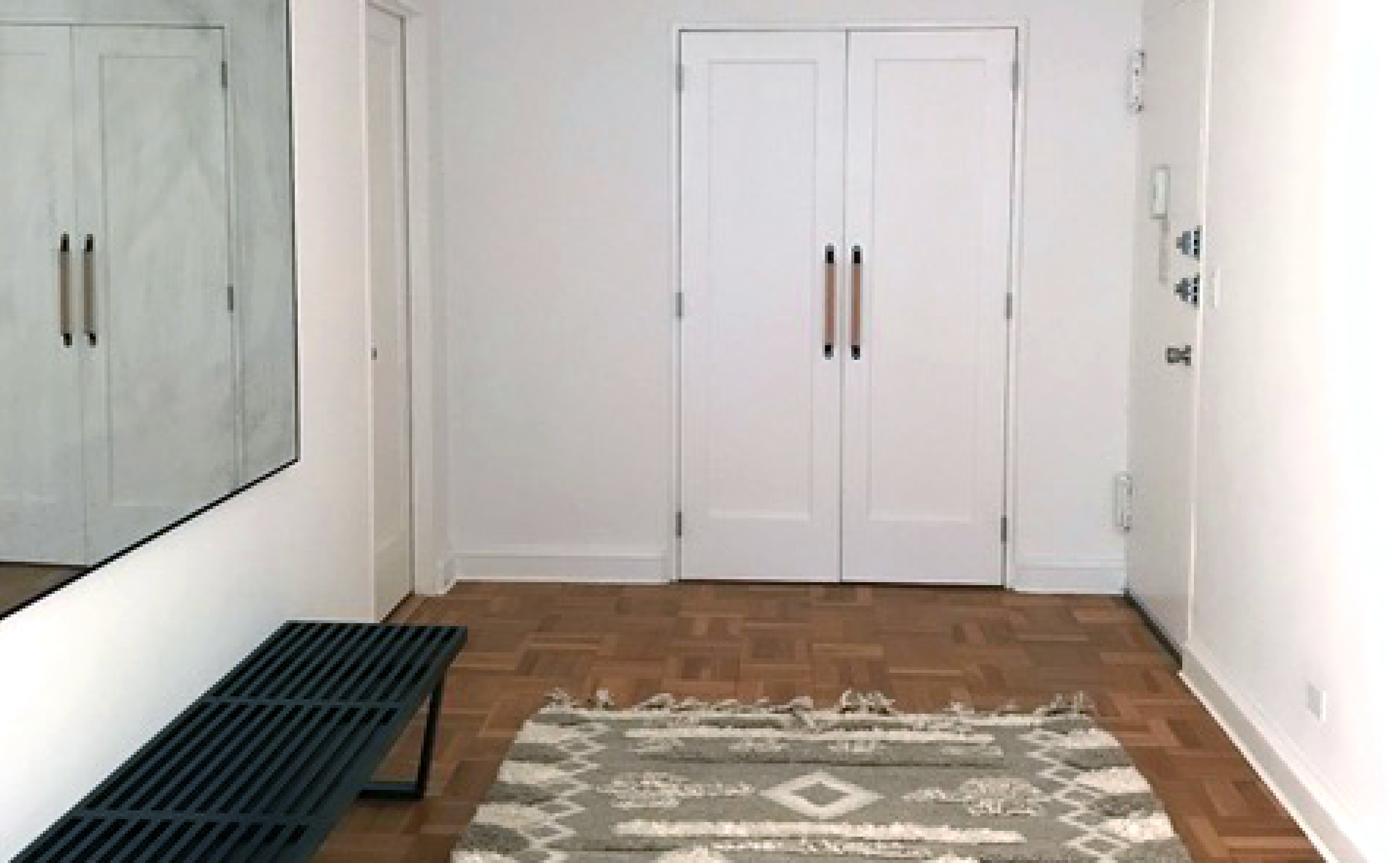 A wide hallway with wood parquet floors and a set of white closet doors at the end of the hallway with wood and leather Sellwood handles