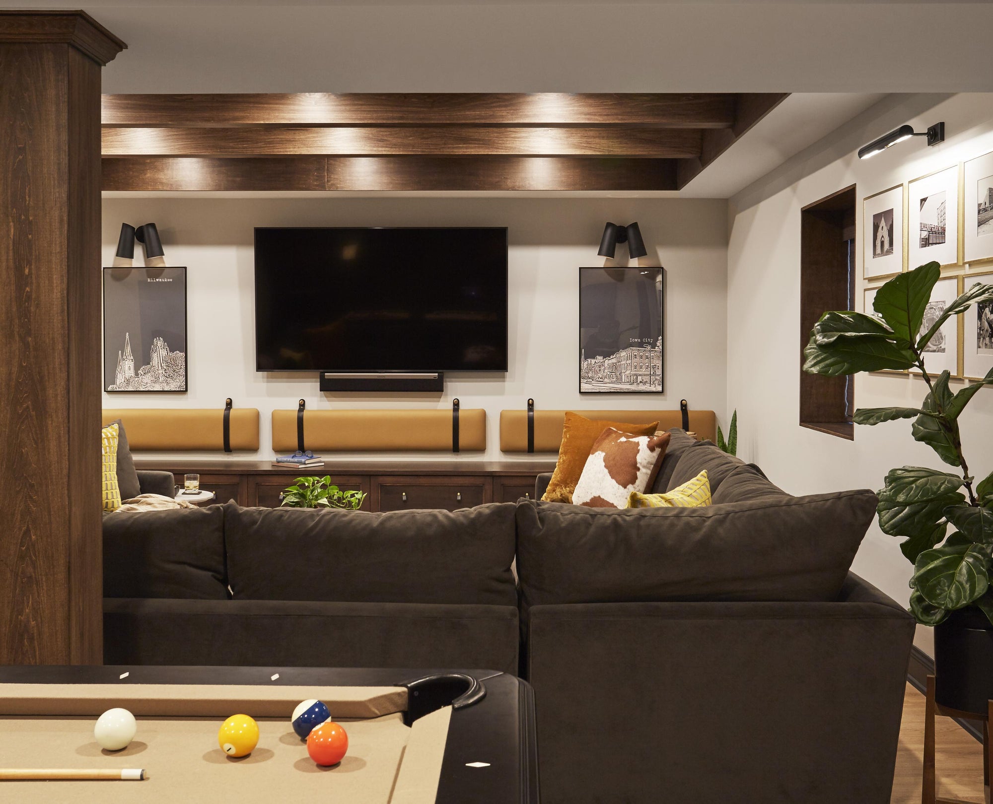 The basement room has a unique ceiling with wood panels, wood beams, and light fixtures inbetween a recessed area to give the illusion of more space, rich brown woodwork and couches, a tan billiards table, and yellow and black leather bench seat