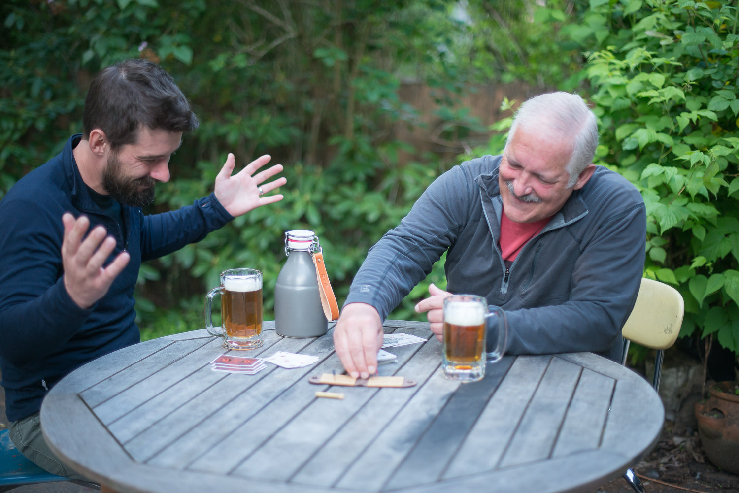 A man and his father are playing cribbage on a picnic table outdoors with two mugs of beer. They are gesturing wildly as one person protests and the other pegs points, they are having fun.