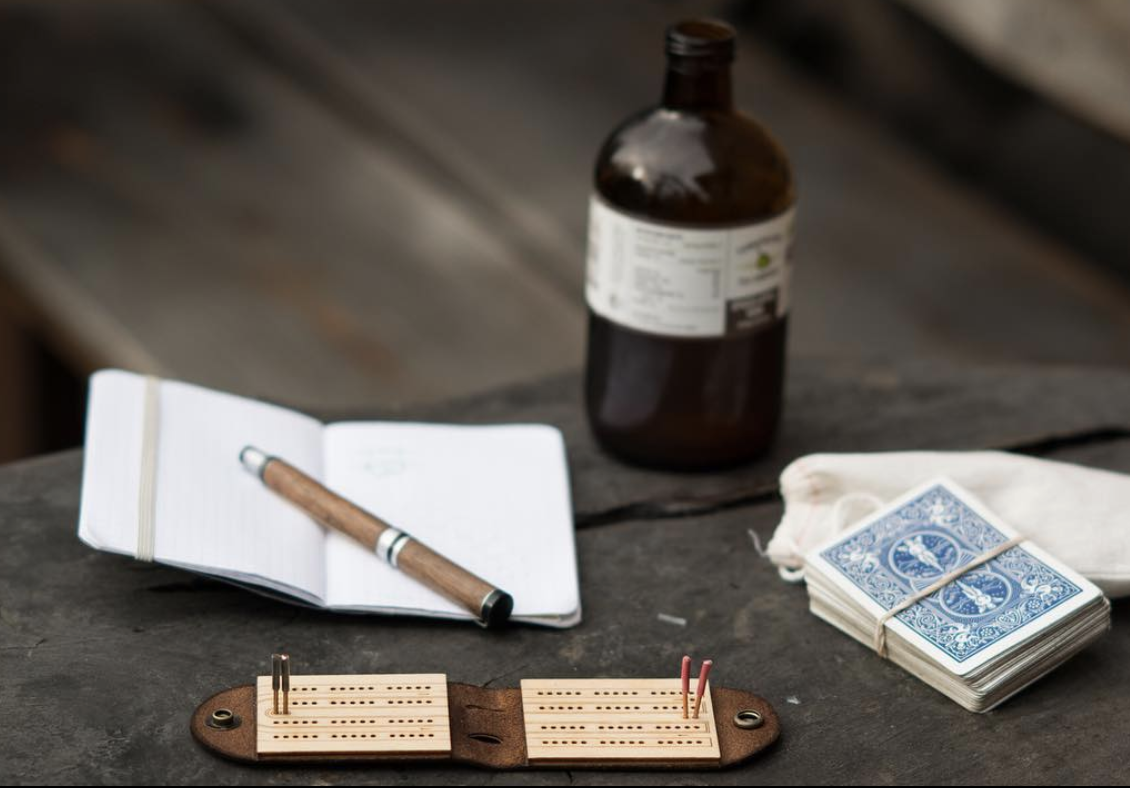 A folding travel cribbage board on a cafe table with a deck of cards and a bottle of kombucha