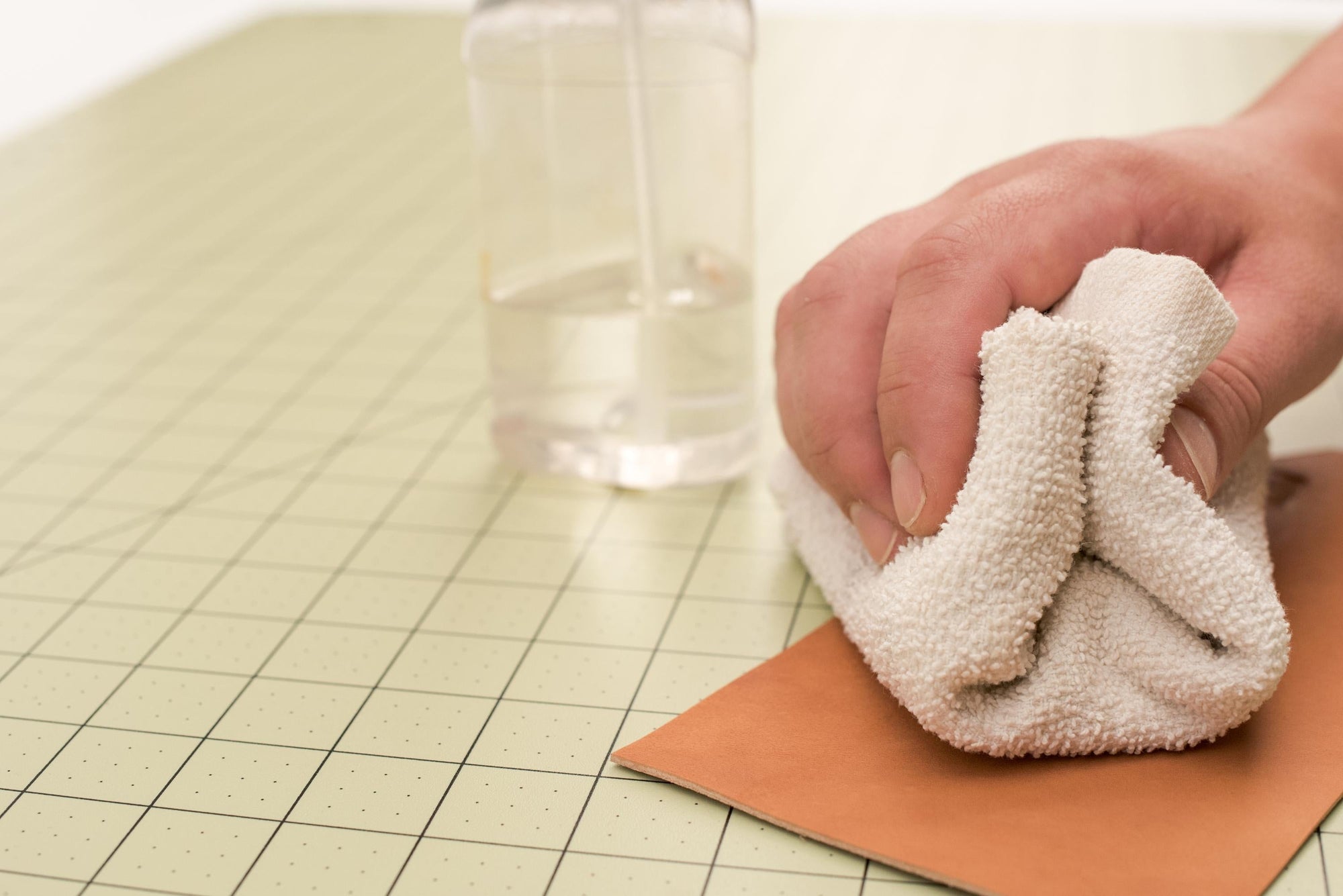 A hand wiping vegetable-tanned leather with a bar towel on a clean surface next to a spray bottle of clear liquid