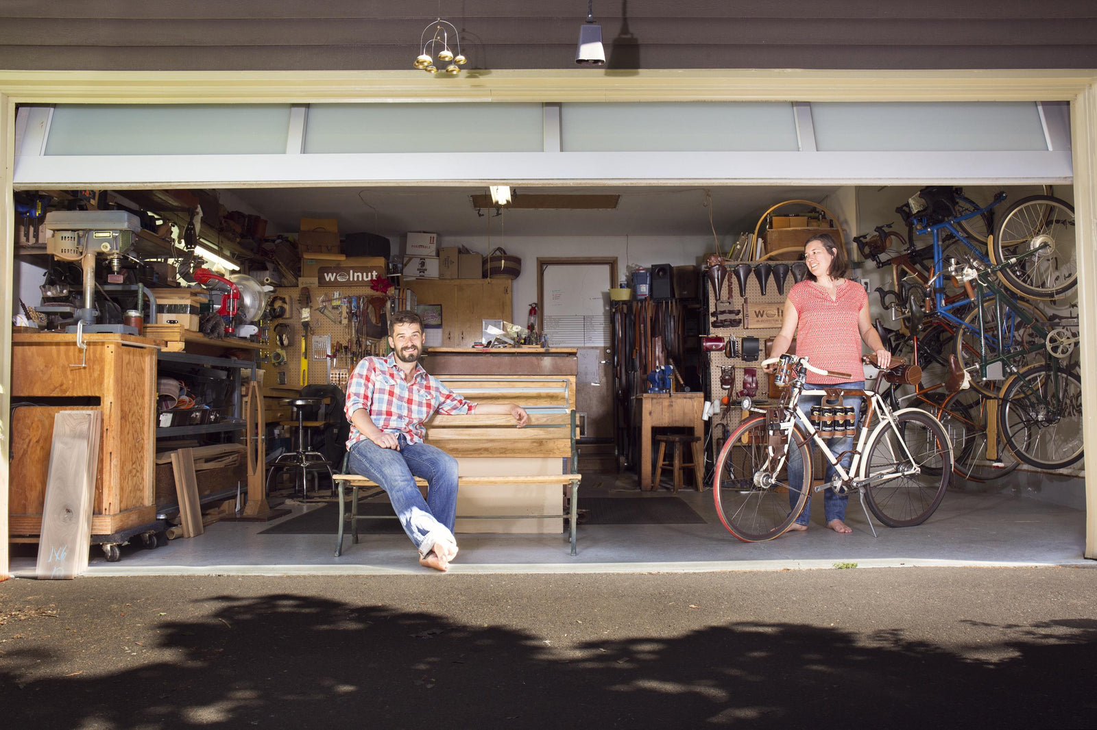 Looking at a garage from the driveway with the door open; Geoff is sitting on a bench in the front smiling; off to the right Valerie is standing with  a bike, and the garage is full of tables, equipment, tools, and benches