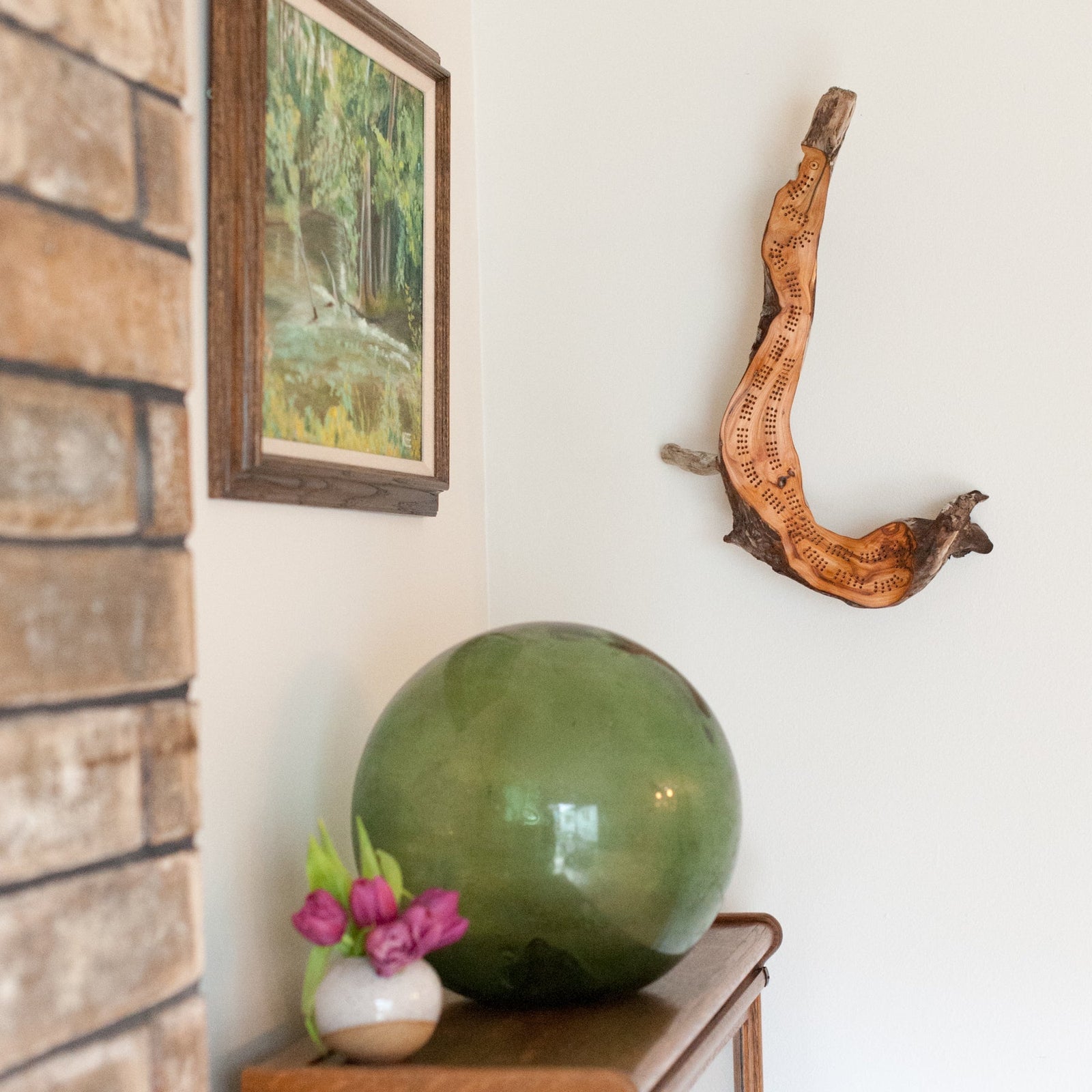 A curved natural driftwood  cribbage board hanging on a white wall surrounded by objets d'art like a sea glass buoy and landscape painting next to a wood shelf and brick wall