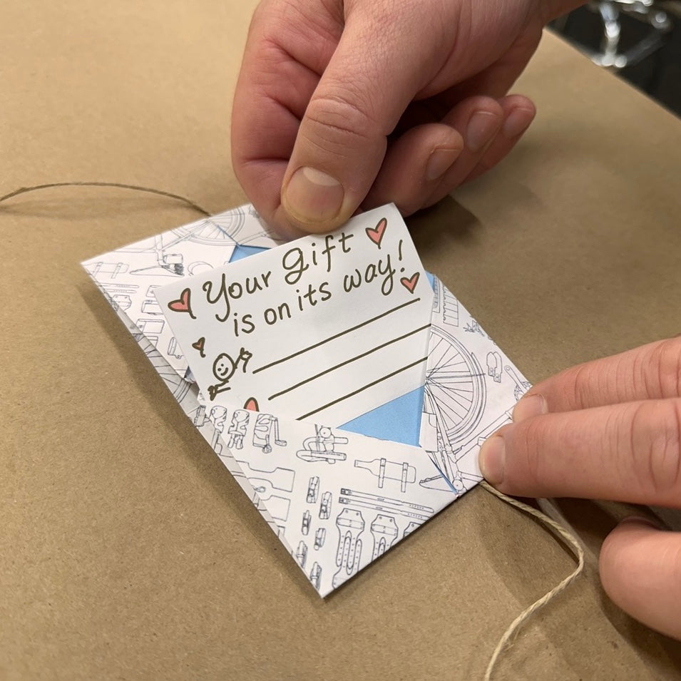 A man is pulling a card out of an origami-folded envelope that says "Your gift is on its way" in handwriting with little heart doodles and hugs