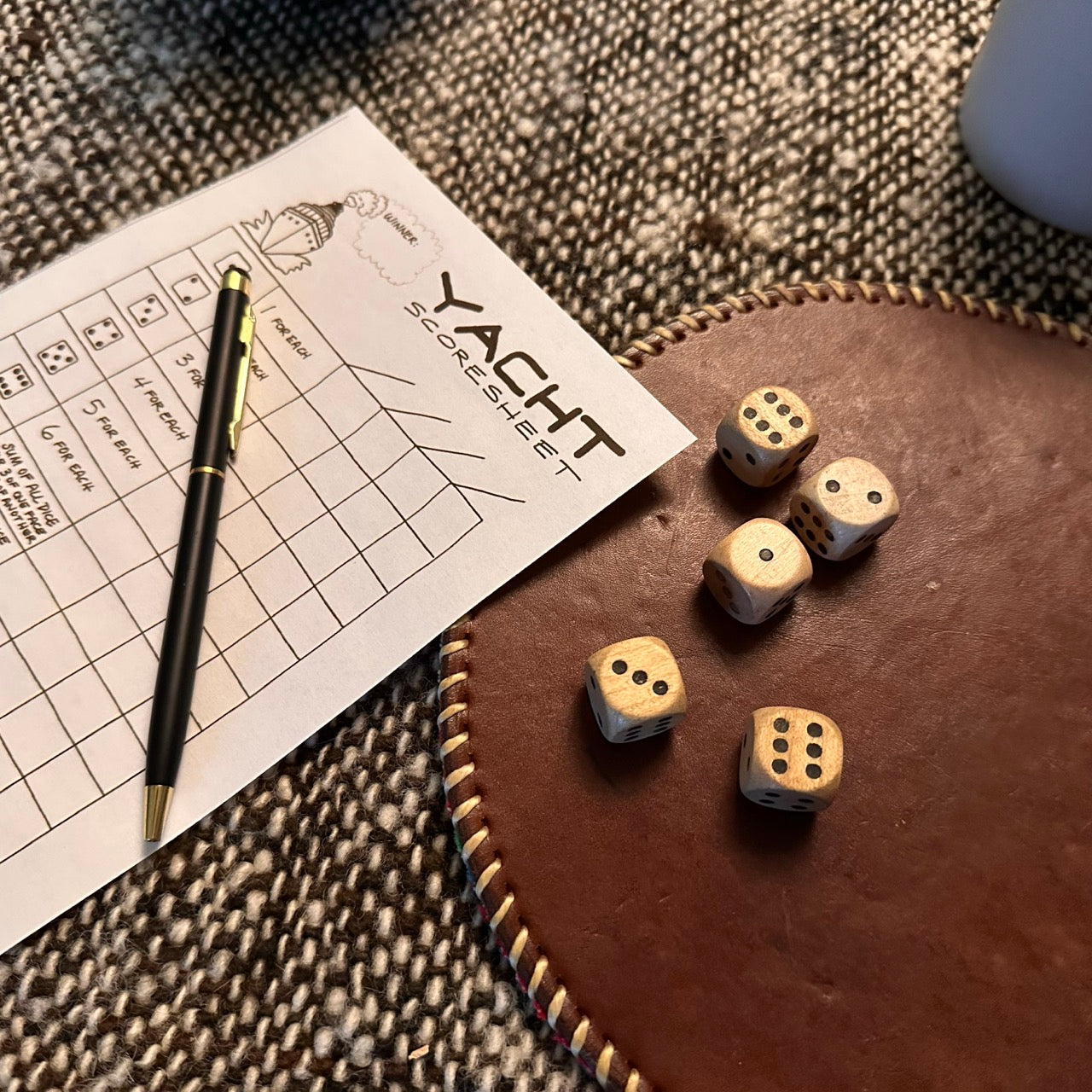 Five wood dice on a leather game playing surface next to a Yacht (Yatzy) scoresheet with a pen on top of a wooly natural textured cloth