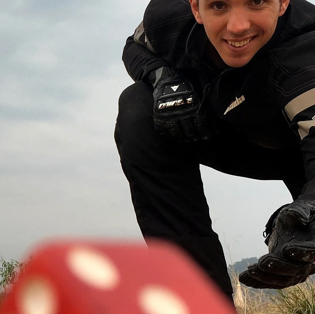 A man wearing black motorcycle gear is kneeling on the ground with his hand outstretched from throwing the dice to decide on his travel destination; the red die he threw is in the foreground.