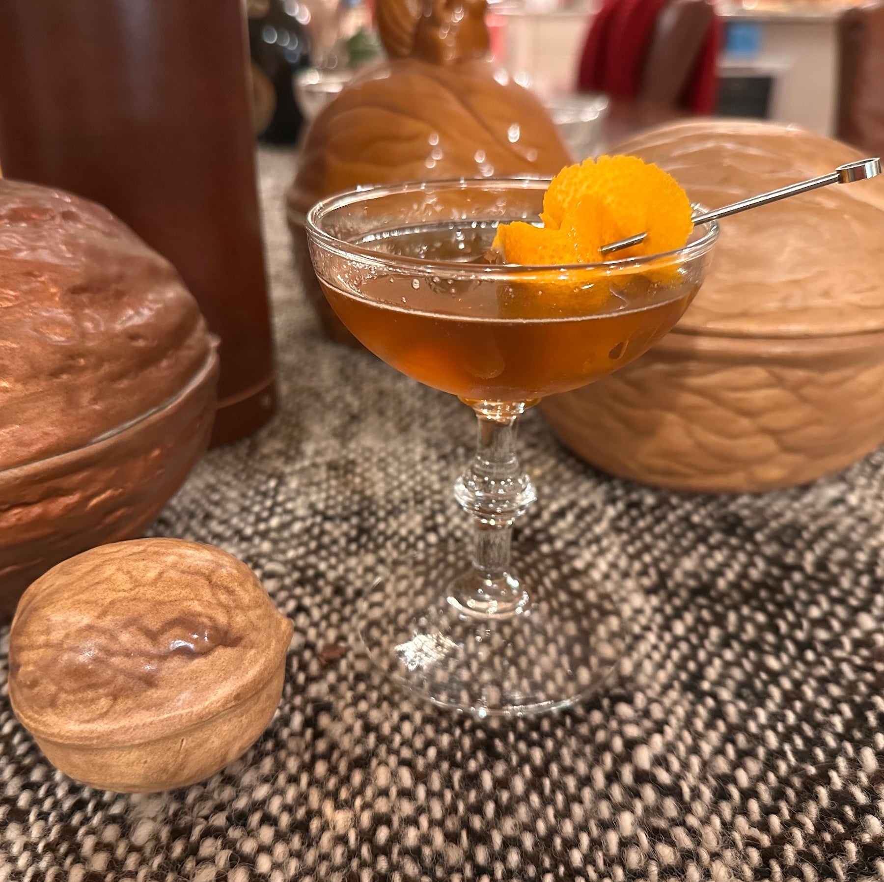 A coupe cocktail glass filled halfway with a cold amber brown liquid and with a curled orange peel on a skewer in front of a collectin of ceramic walnuts on a dark brown and white wool tablecloth,and a leather whiskey tube in the background