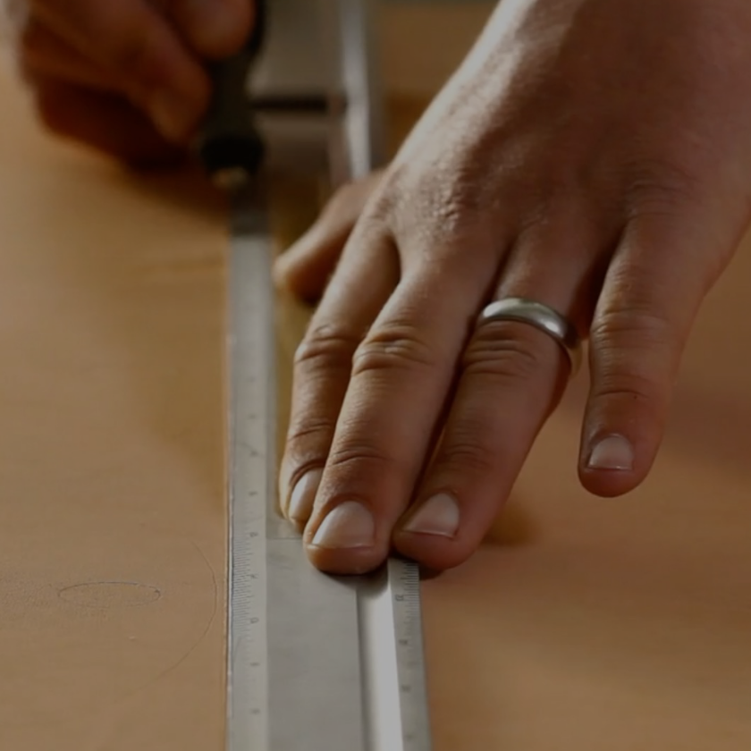 A leather craftsman's hand with a wedding ring on holding down an aluminum ruler on a veg-tan leather hide and cutting with his right hand using an exacto blade