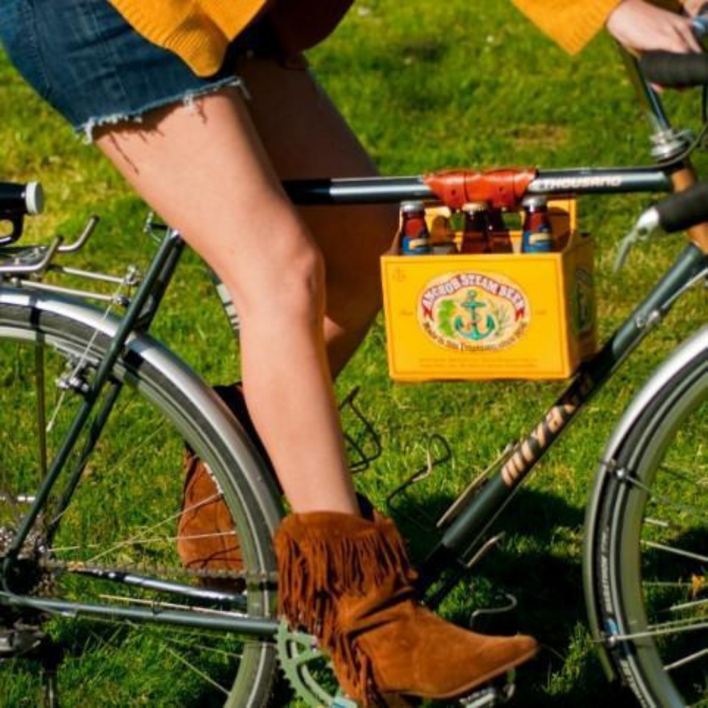 Woman cycling on a sunny grassy lawn with a 6-pack of yellow Anchor Steam beer strapped to her top tube with a honey leather carrier strap