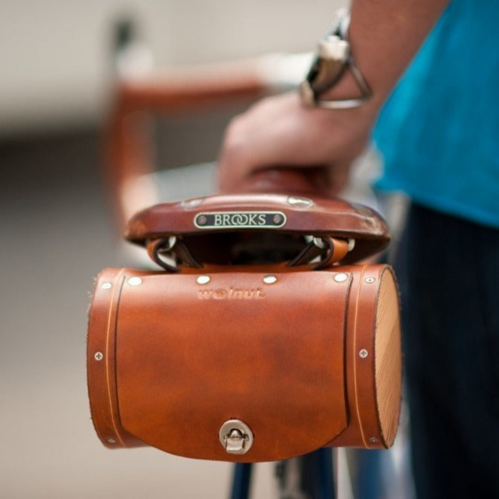 Close-up of a honey leather barrel bag mounted to the back of a Brooks bicycle saddle, with a man in a turquoise t-shirt resting his hand on the saddle and looking down the bike towards matching honey leather bar wraps