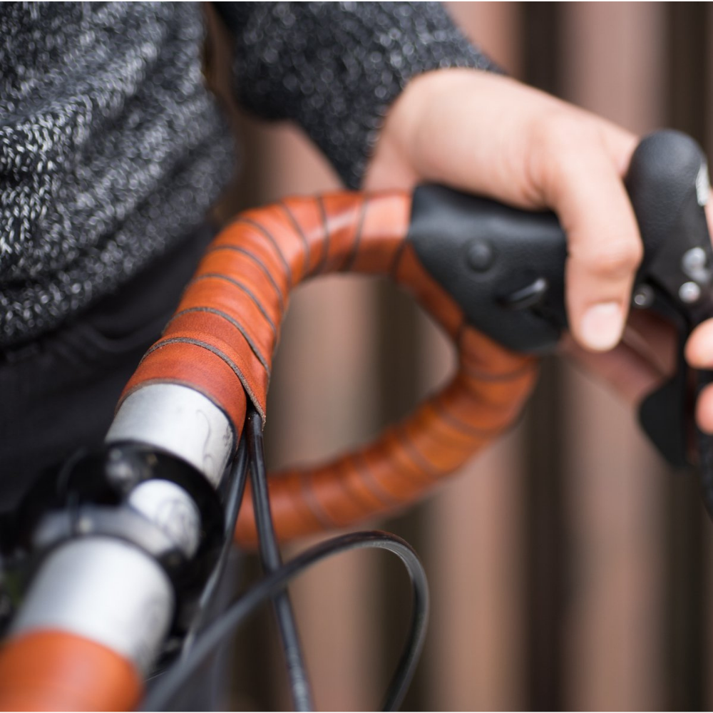 Close-up of traditional bicycle handlebar drop bars wrapped in a lush and tactile honey brown leather, held by a person whose hips and hand are visible against an urban rusty background