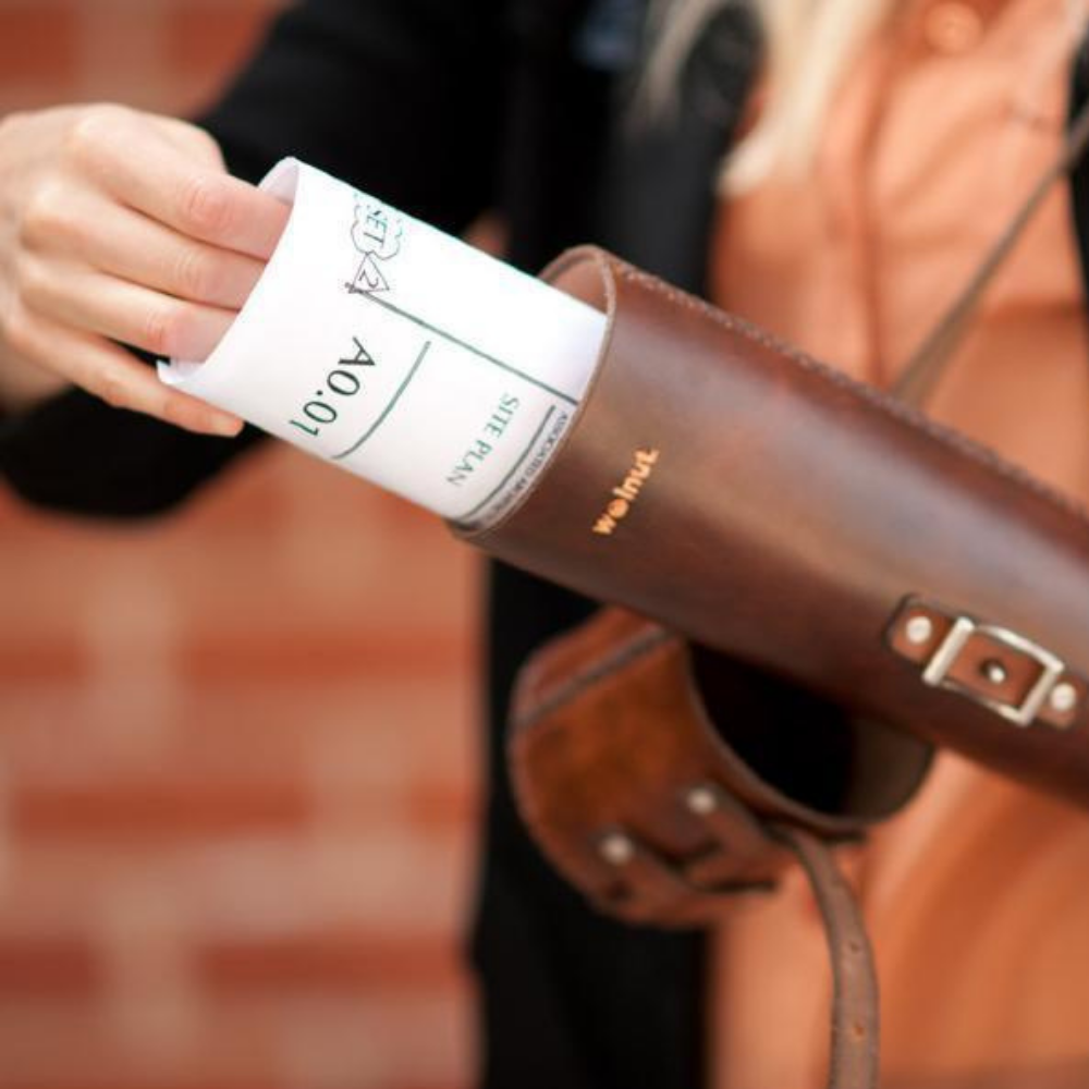 A woman in front on an urban city brick wall pulling a set of architectural drawing plans out of a dark brown leather document tube