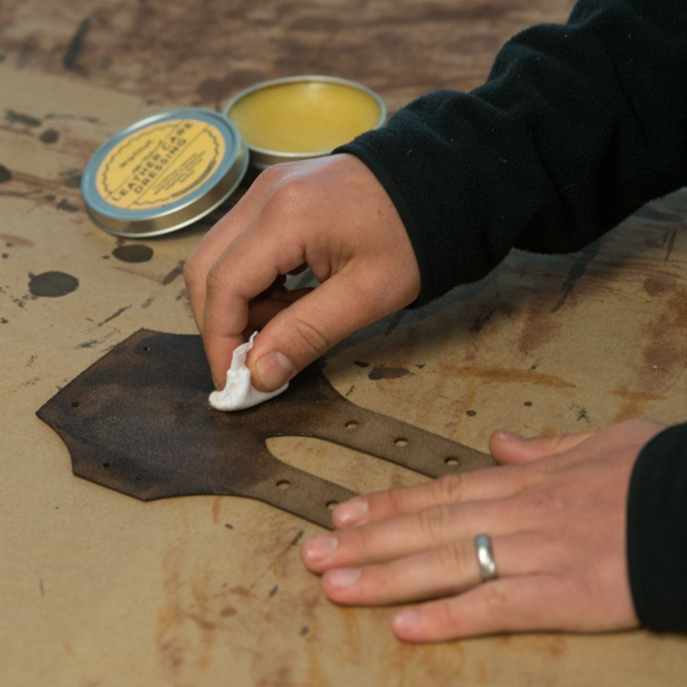 Close-up of hands rubbing in conditioner to a piece of leather on a stained work surface using a clean white rag, with a tin of Leather Care in the background
