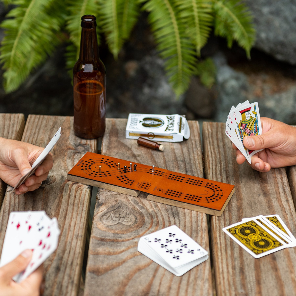 Three people playing card games outside on a wood picnic table in front of ferns and rocks on a leather cribbage board