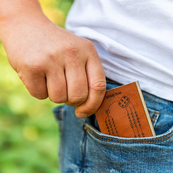 A man's hand sliding a small leather game into his denim blue jeans pocket outdoors against greenery