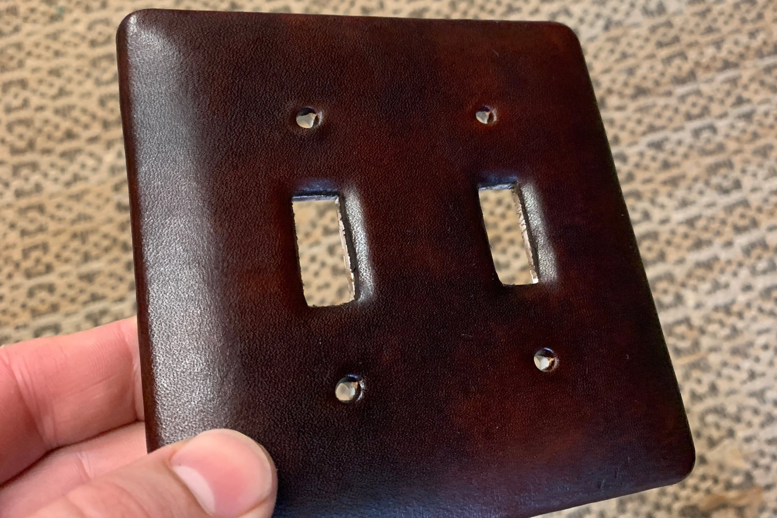 Close-up of a hand holding a light switch plate that is covered in dark brown leather, a custom project for a residential new build fabricated in our workshop