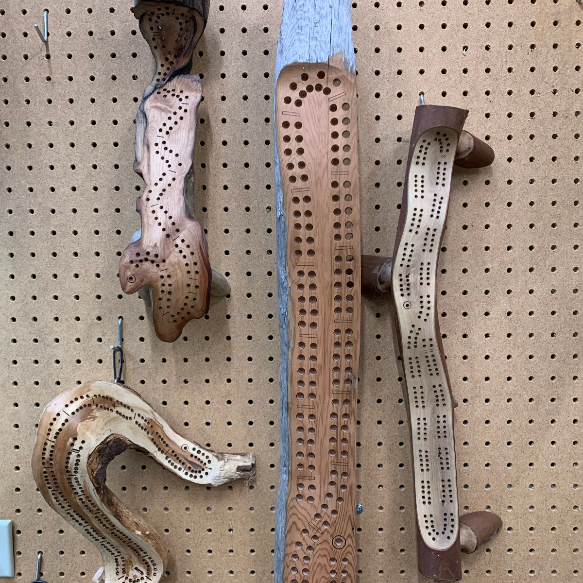 A variety of shaped- and sized- driftwood  cribbage boards hanging on a pegboard