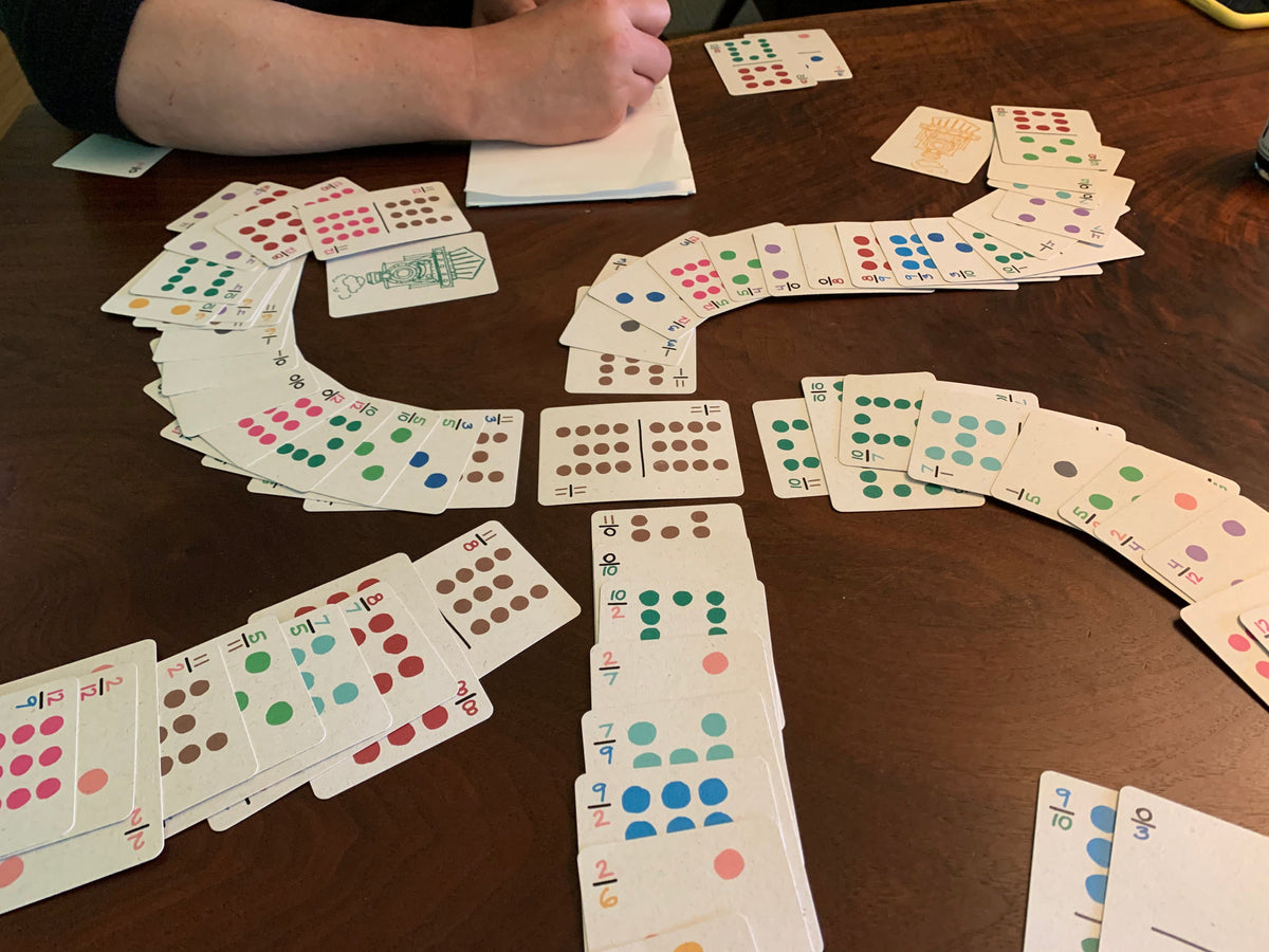 A close-up of a game board made of playing cards spread out in domino trains from a central hub on a wooden table, with a person's arm and hand visible writing on a small notepad. The card trains are compressed because only the index corners are visible. 