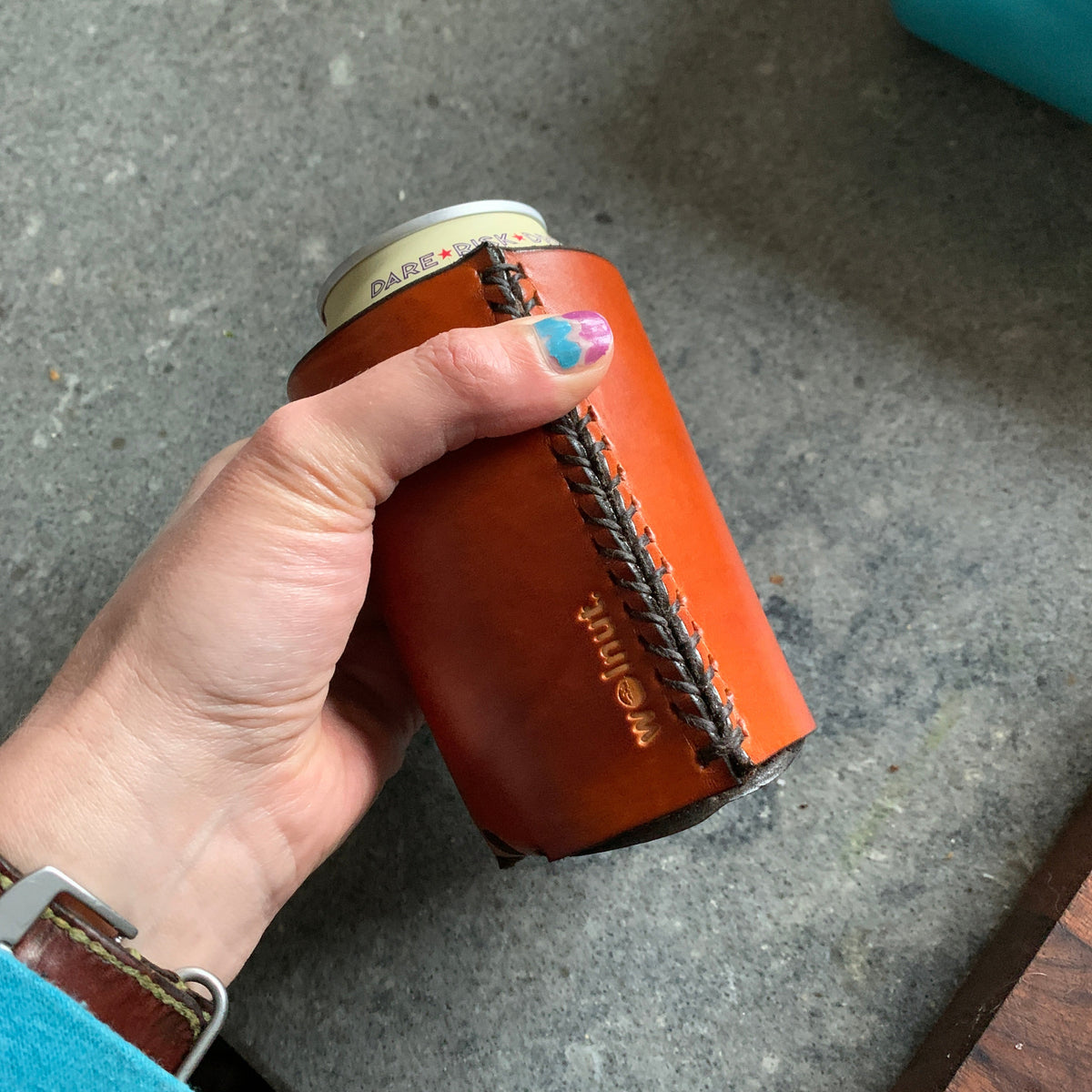 Hand holding a honey brown leather can sleeve (cookie) with 'Walnut' branding on a concrete surface.