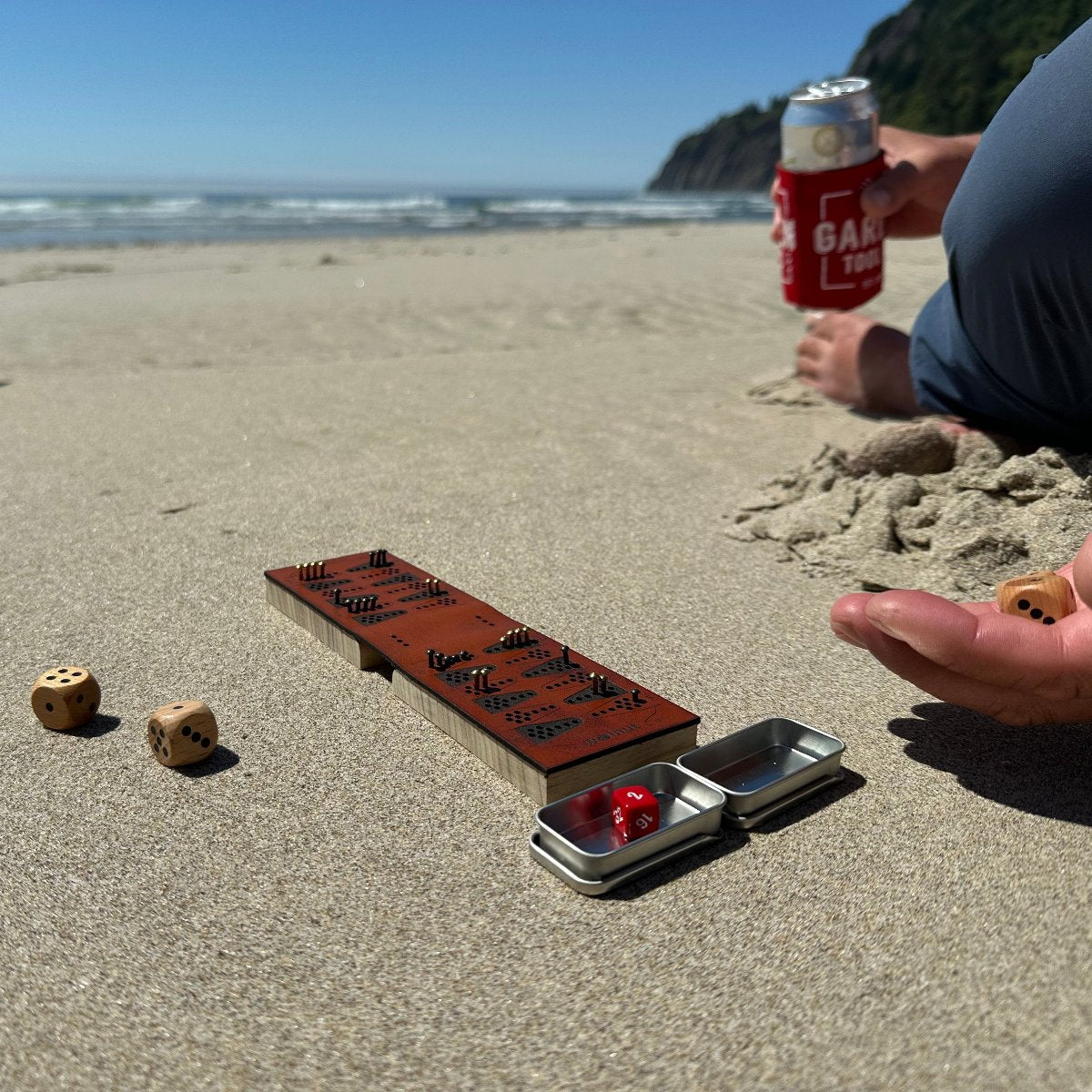 Photo taken at the beach, with a travel backgammon board on the sand, the camera facing the ocean. A person is sitting to the right side mostly off-camera, rolling dice in his hand.A relaxing way to play backgammon on the go - &quot;beachgammon&quot;?