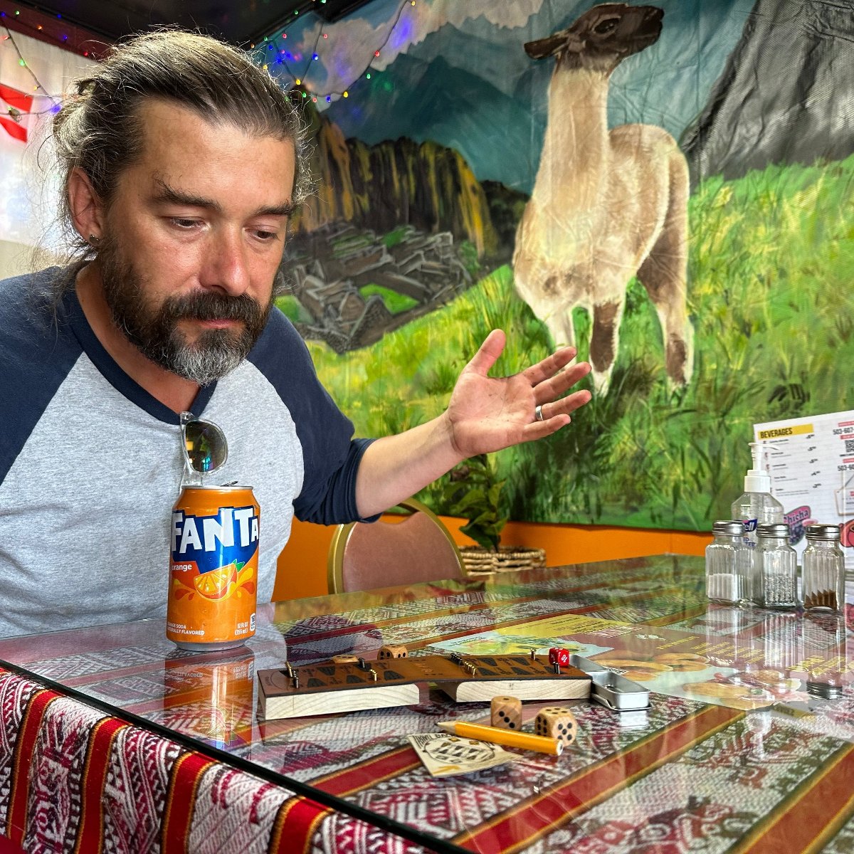 A man is playing travel backgammon in a Peruvian restaurant, with a mural of a llama in the Andes on the wall behind him. On the restaurant table surface is the small leather backgammon board and a Fanta orange can of soda. He is rolling the dice and staring at the board intently. 