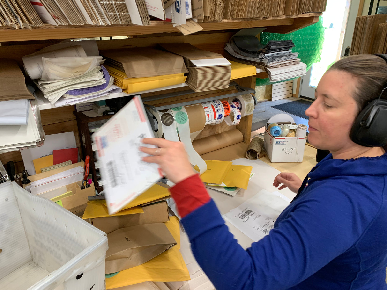 A woman is busy at work at a shipping and packaging station, her hand is blurred as she moves a Priority mail envelope to a USPS mail bin