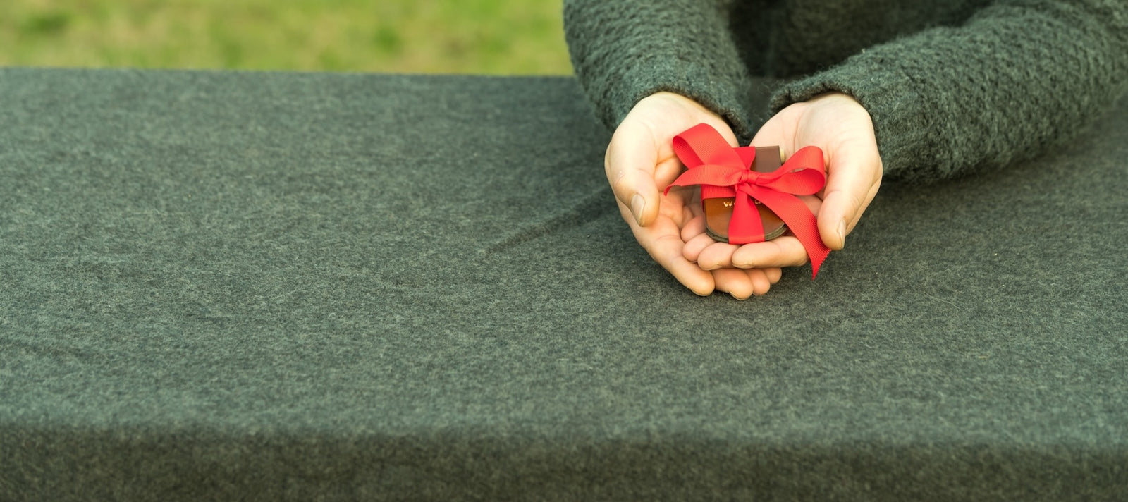 A man in a green sweater resting his arms on a green wool army blanket and holding out a small wrapped leather cribbage board in his hands, which make the shape of a heart. The board is a wrapped in a red gift ribbon. 