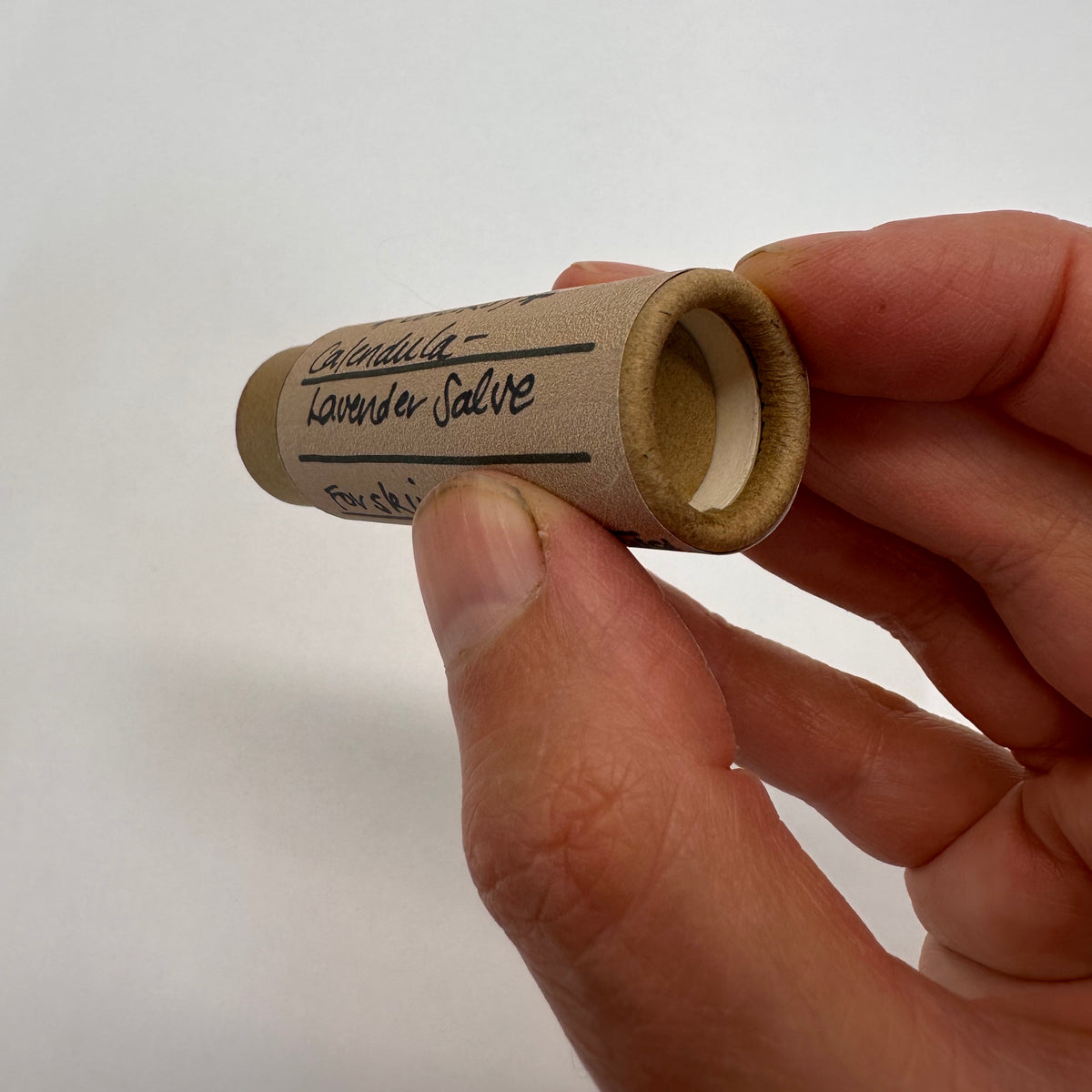 Hand holding a small cardboard tube labeled &#39;Lavender Salve&#39; against a white background showing the bottom where the finger will push up