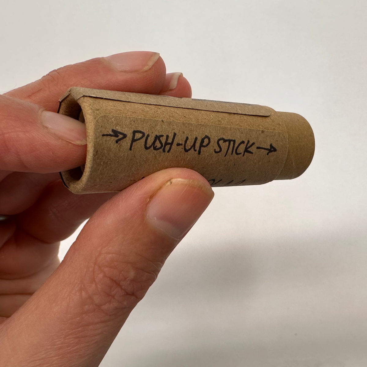 Hand holding a cardboard tube labeled &#39;Push-up Stick&#39; against a plain background with the finger pushing up to dispense