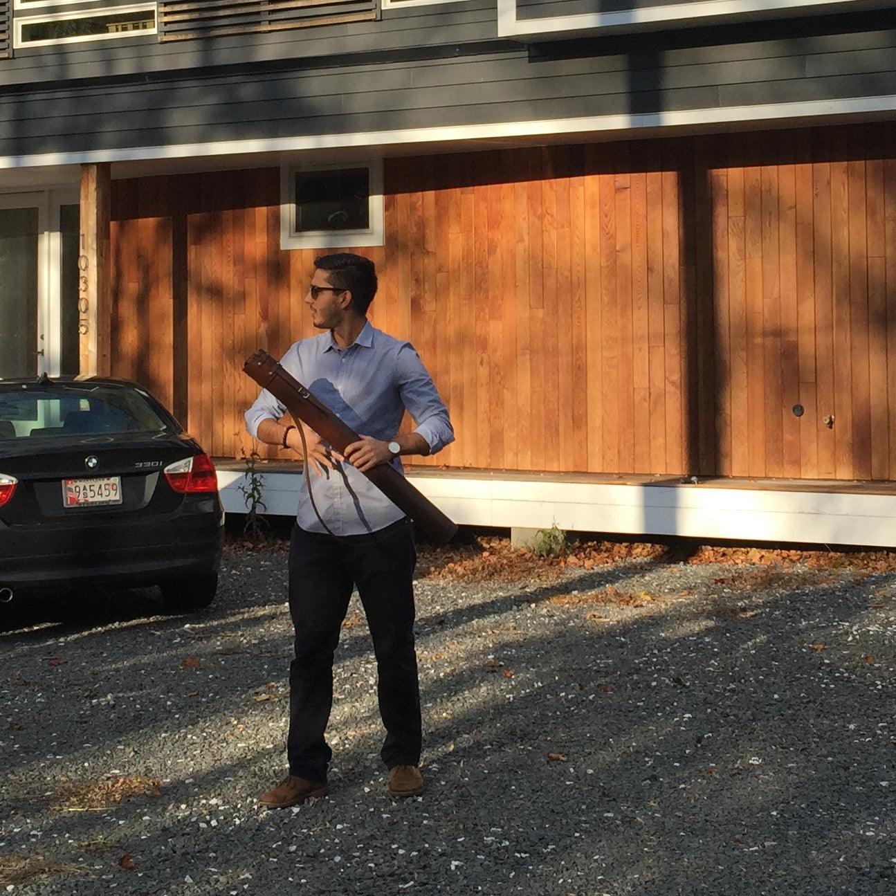 An architect is walking away from a newly-built house through the gravel driveway, while fastening his dark brown leather blueprint plan document case and looking off to the right. 