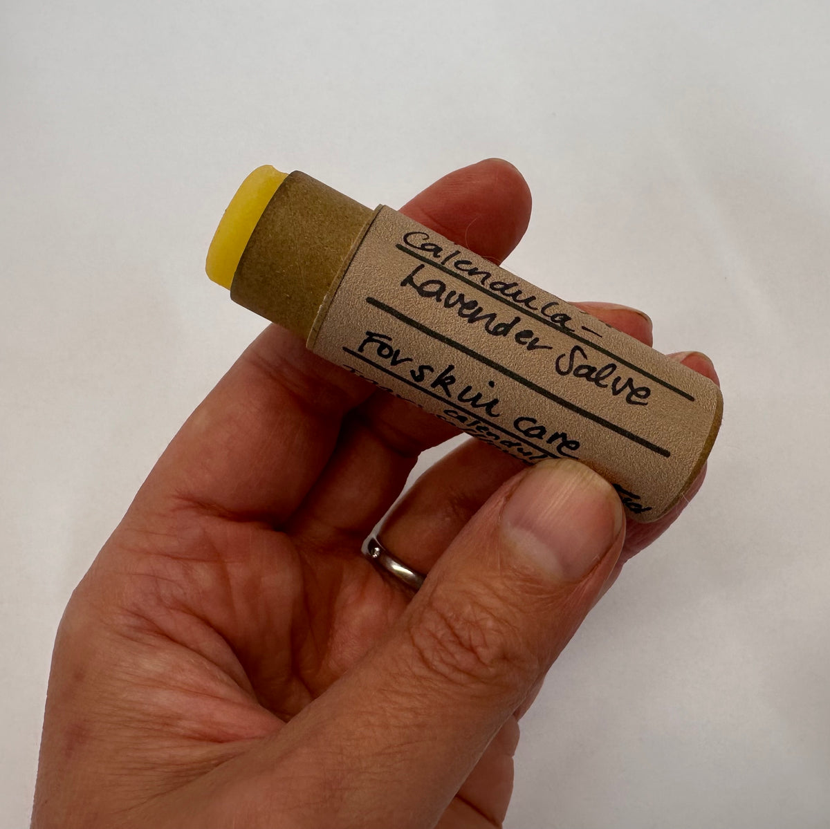 Hand holding a calendula lavender salve stick on a white background, the orange-ish rich salve is pushed out of the tube