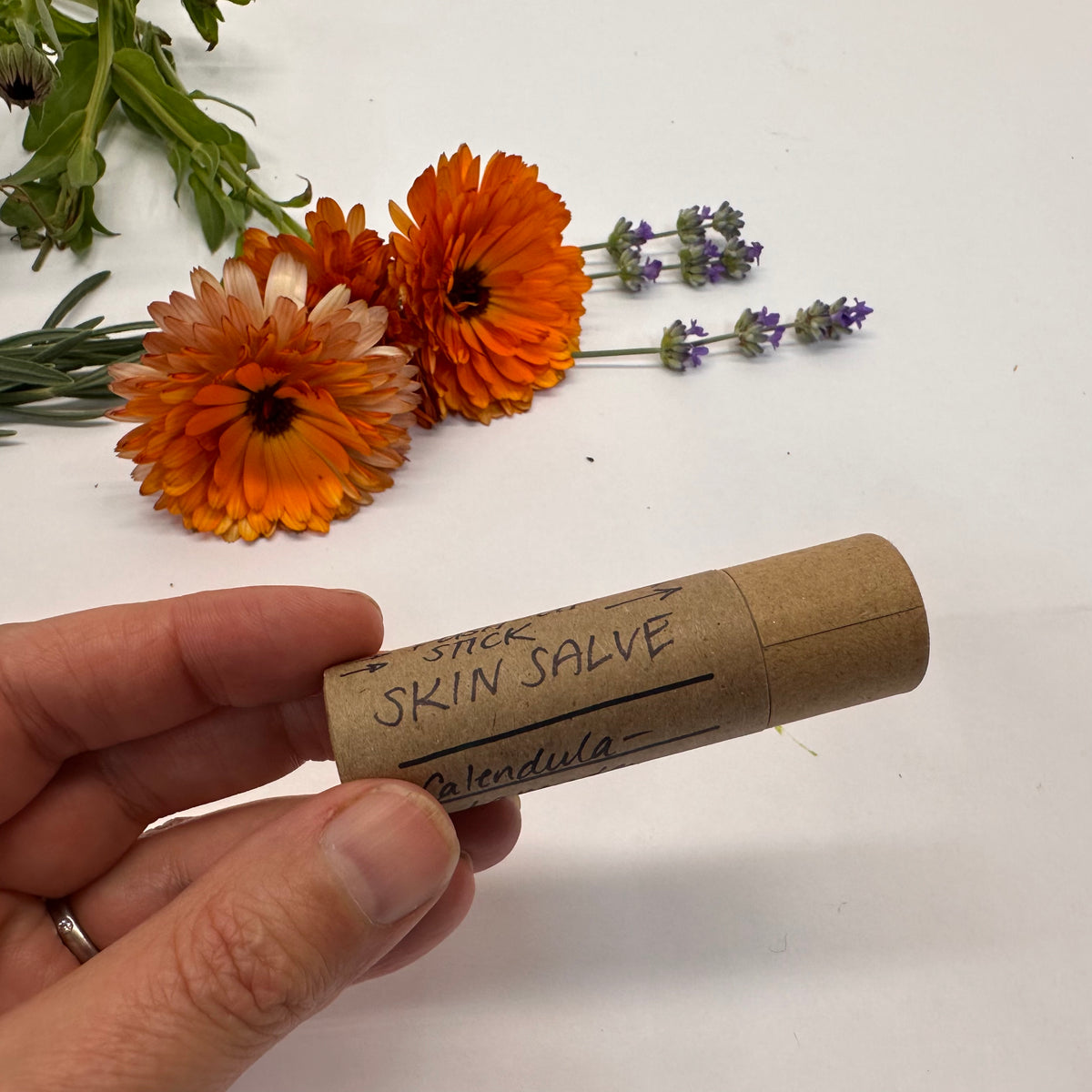 Hand holding a skin salve stick labeled &#39;Calendula&#39; with flowers in the background.