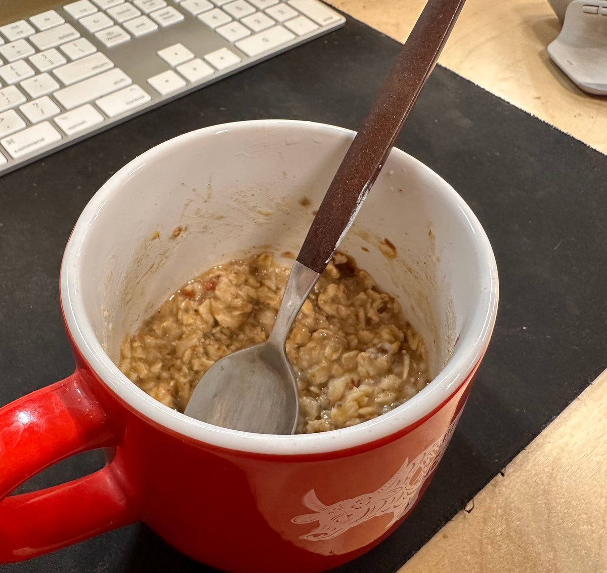 Red mug with oatmeal and a spoon on a desk in front of a keyboard and mouse