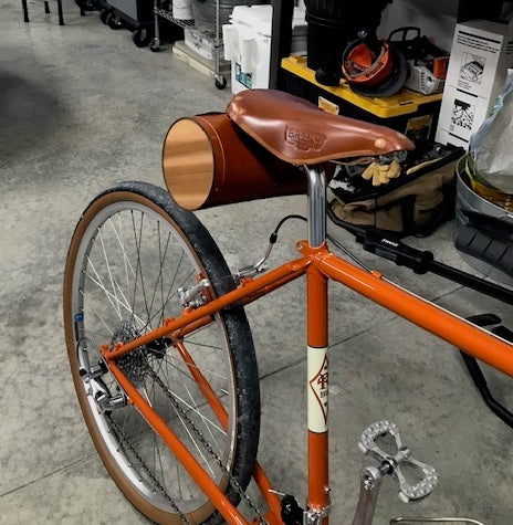 Customer photo of an orange bicycle with a dark brown Brooks leather saddle and honey XL leather barrel bag in a garage