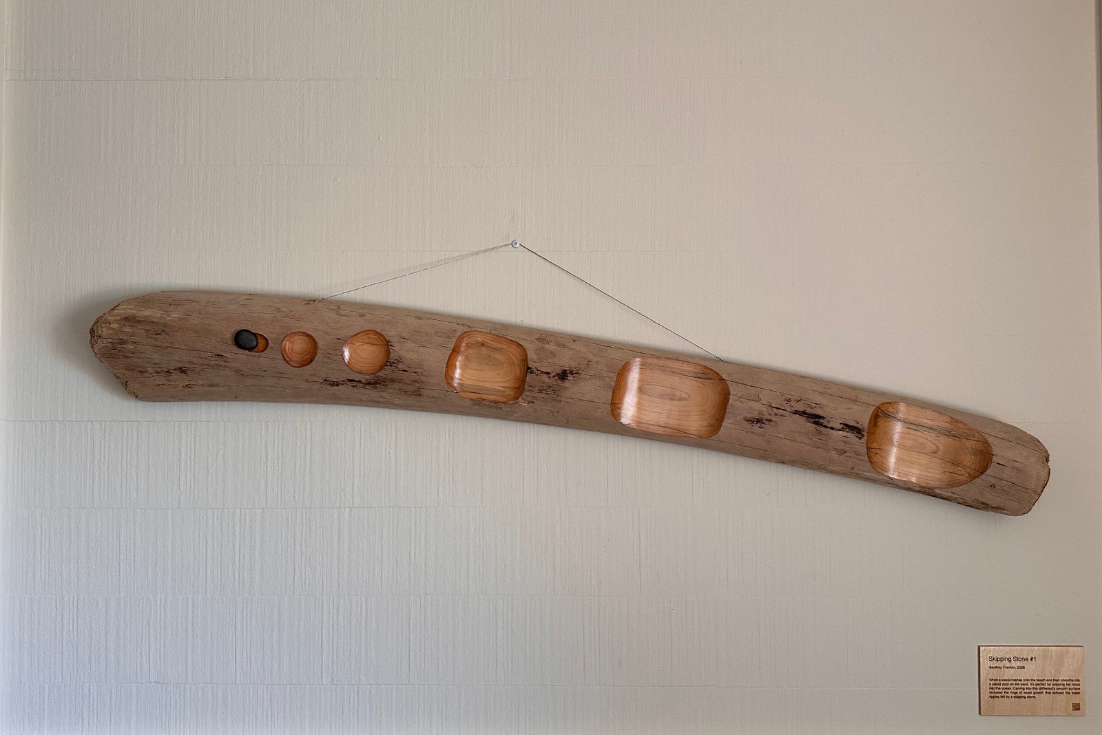 Driftwood art piece with carved away surface dots and a skipping stone on a white wall