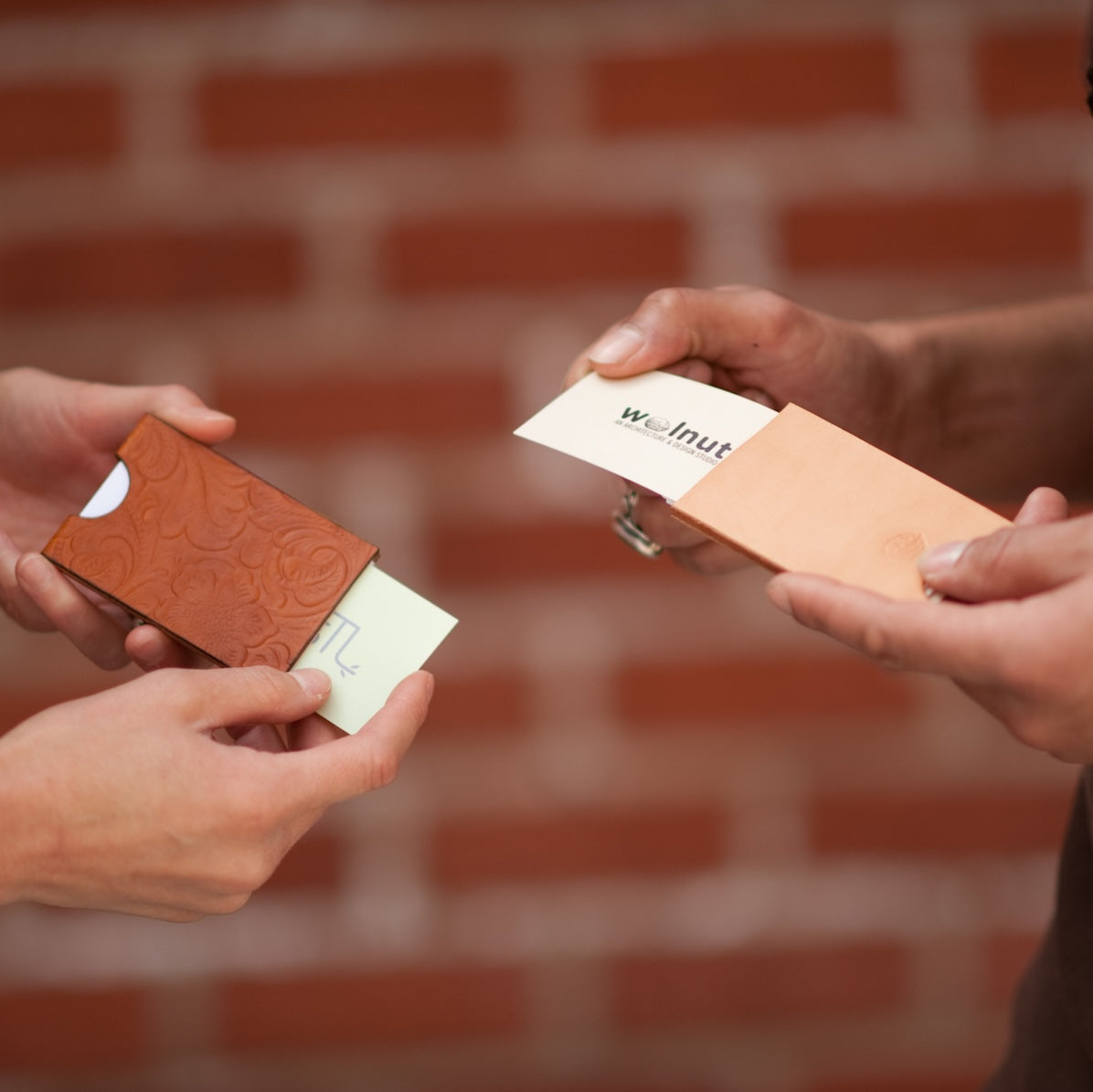 Two people exchanging business cards that are in each leather business card cases, in front of a brick wall