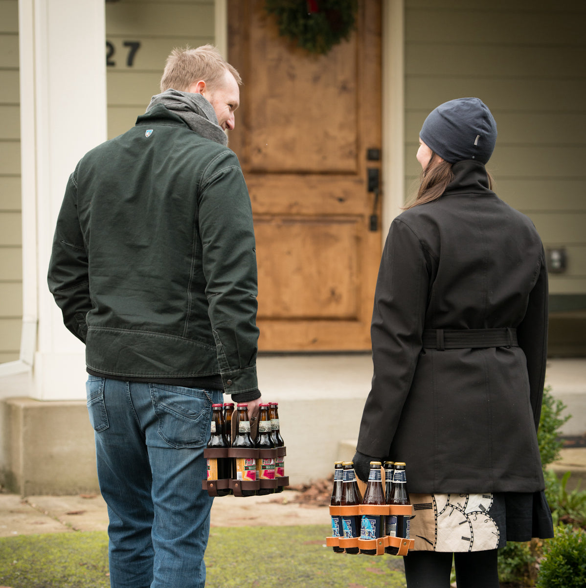 Two friends, a man and a woman, are walking to the front door of a house, each carrying a leather and wood 6-pack with brown glass beer bottles. 