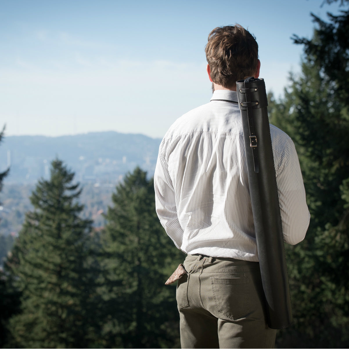 A man standing in the Pacific Northwest at a park, looking off into the distance at a mountain vista between large Doug Fir trees, with a black leather blueprint document case slung over his shoulder