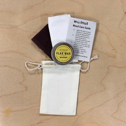 A small wood care finish kit displayed on a light wood tabletop, including a 1-oz circular metal tin with a yellow label "Flax Wax", a white Wood Care Guide how-tocard, an abrasive pad, a white buff rag, and a white drawstring pouch.