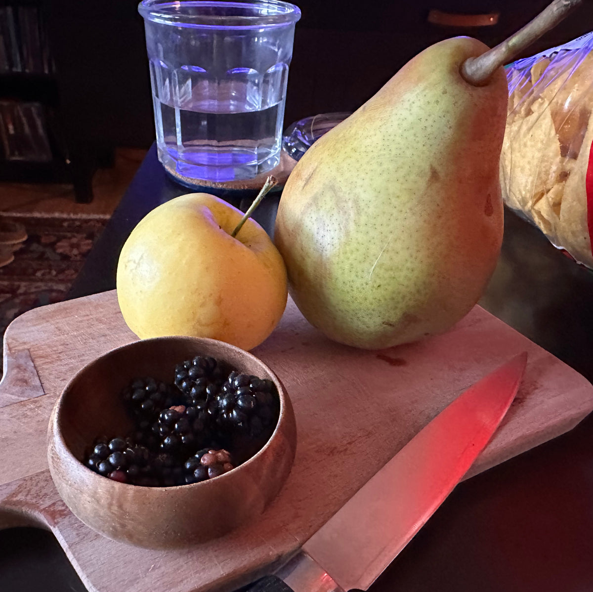 Pear, apple, bowl of blackberries, and knife on a wooden cutting board with a glass of water in the background.