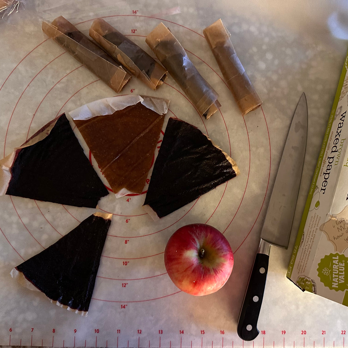 Cutting mat from a bird&#39;s eye view, with wax paper wrapped fruit rolls, cut fruit rolls that are about 1/8 of a dehydrator tray, an apple, and a knife