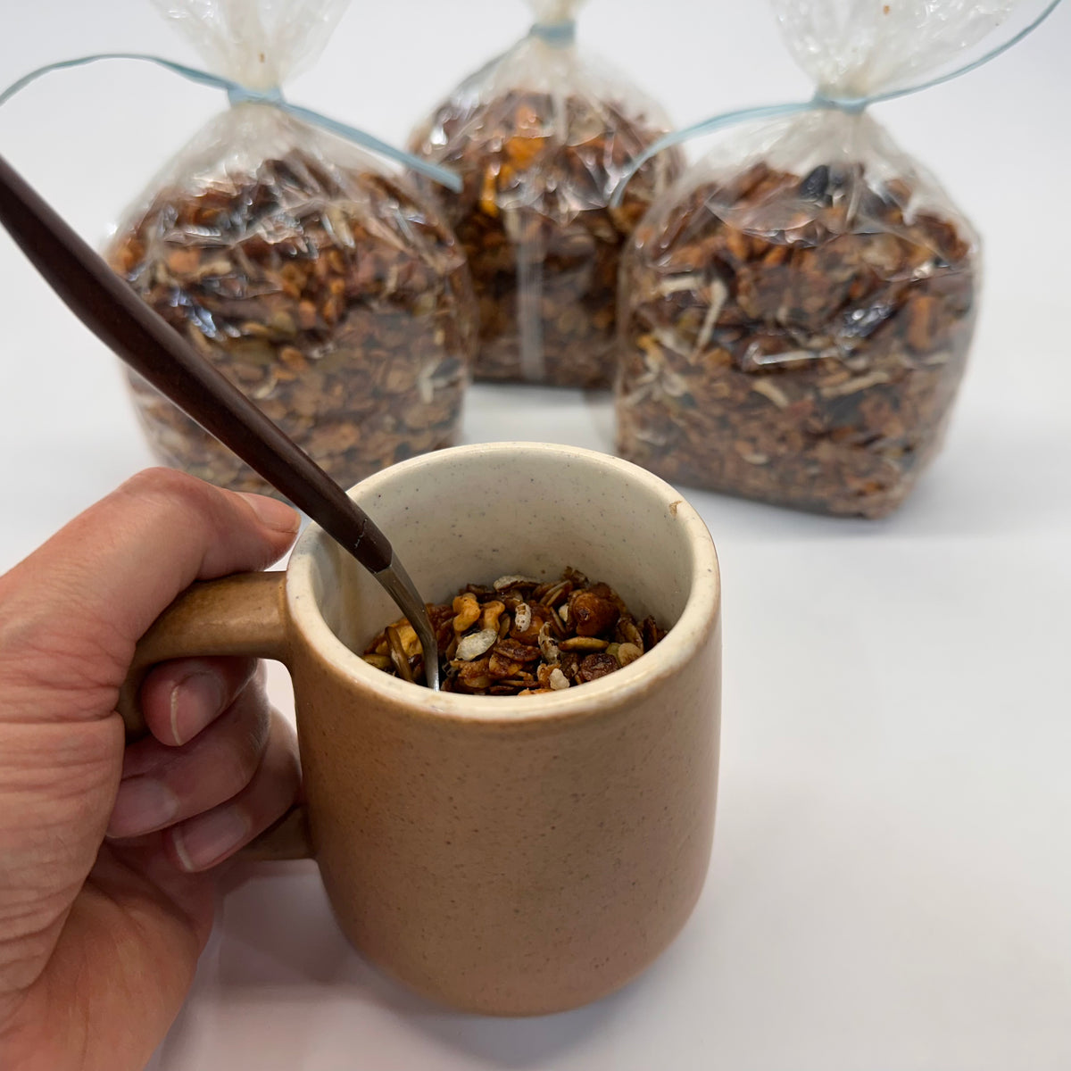 Person holding a brown mug of granola being used as a small bowl with a spoon in it, with bags of granola in the background