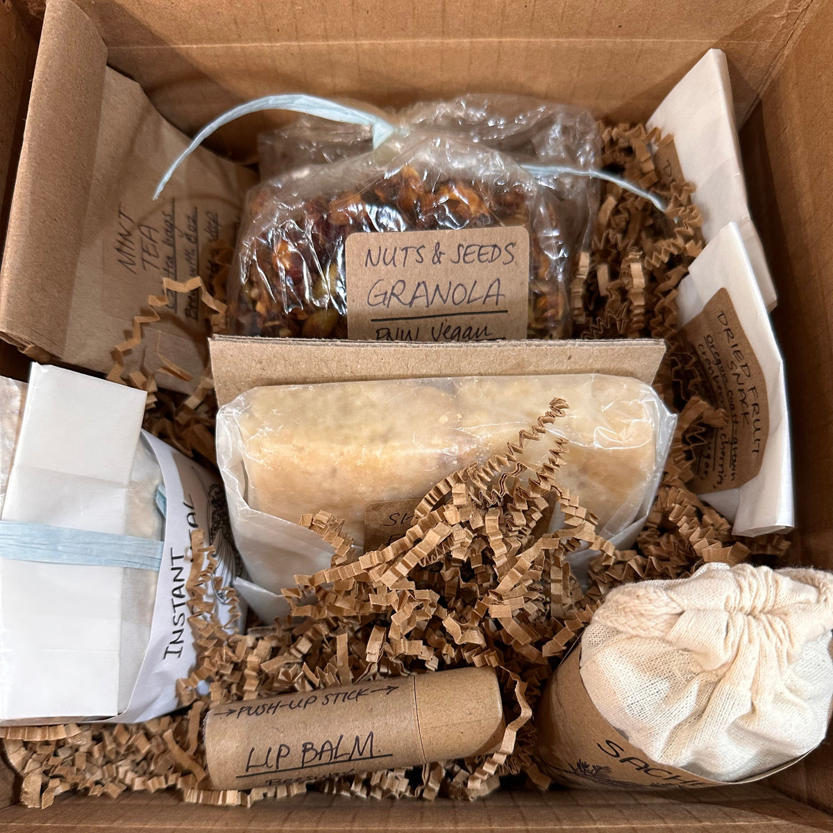 Box containing various items wrapped in natural packaging including products with labels, Nuts and Seeds Granola, Lip Balm, Mint Tea, Sachet, Instant Oats, Dried Fruit Snacks