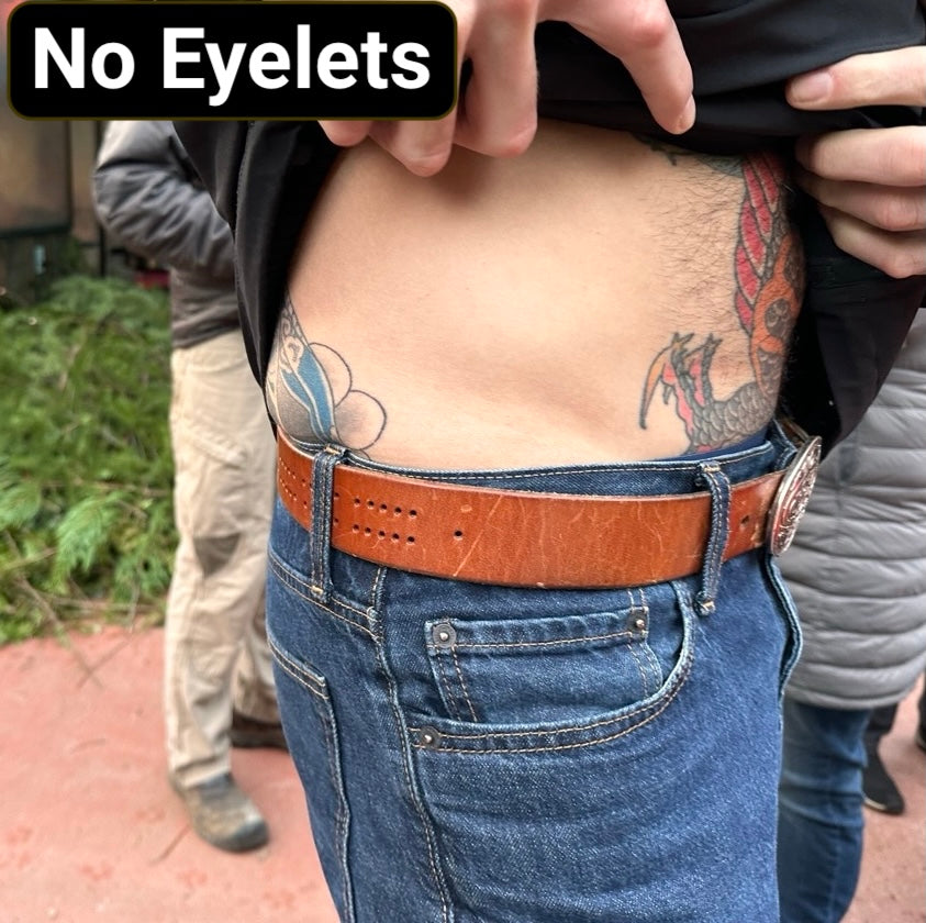Person wearing blue jeans with a light brown cribbage board belt that has a custom oval buckle, with a tattooed torso and text 'No Eyelets' at the top.