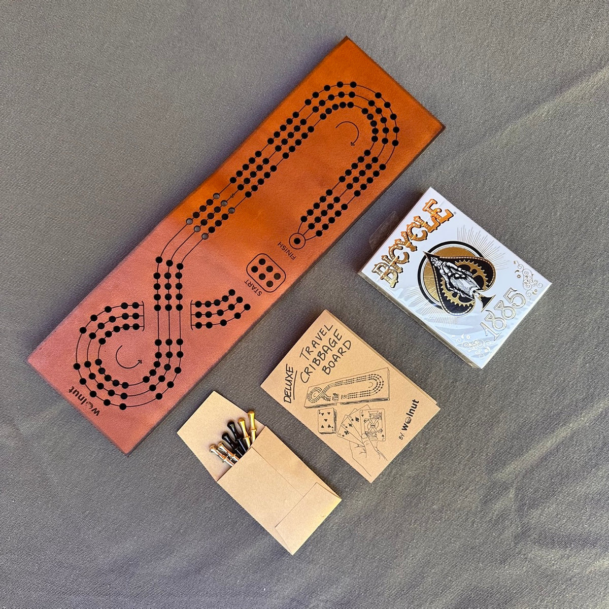The leather travel cribbage board laid out on flat on a grey cloth surface with a small envelope with 3 sets of pegs in nickel, brass, and black and a small paper mini book with rules of play, and a deck of BIcycle brand playing cards in gold and black with the design &quot;1885&quot;