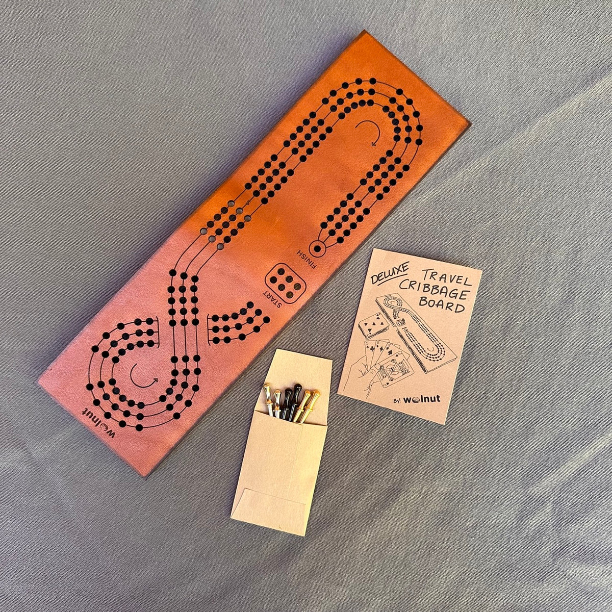 The leather travel cribbage board laid out on flat on a grey cloth surface with a small envelope with 3 sets of pegs in nickel, brass, and black and a small paper mini book with rules of play