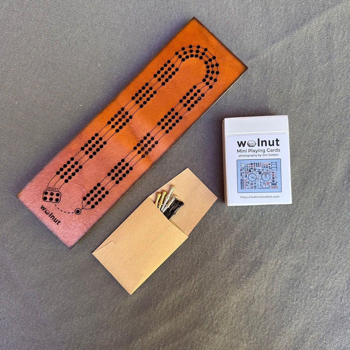 The travel cribbage board laid out on flat on a grey cloth surface with a deck of half-sized playing cards and a small envelope with 3 sets of pegs in nickel, brass, and black
