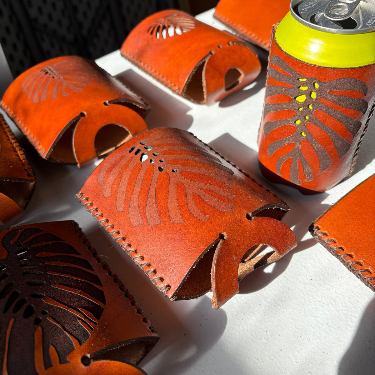 Brown leather can coolers with cut-out designs, lying flat folded and shown with a can, on a sunny market table surface.