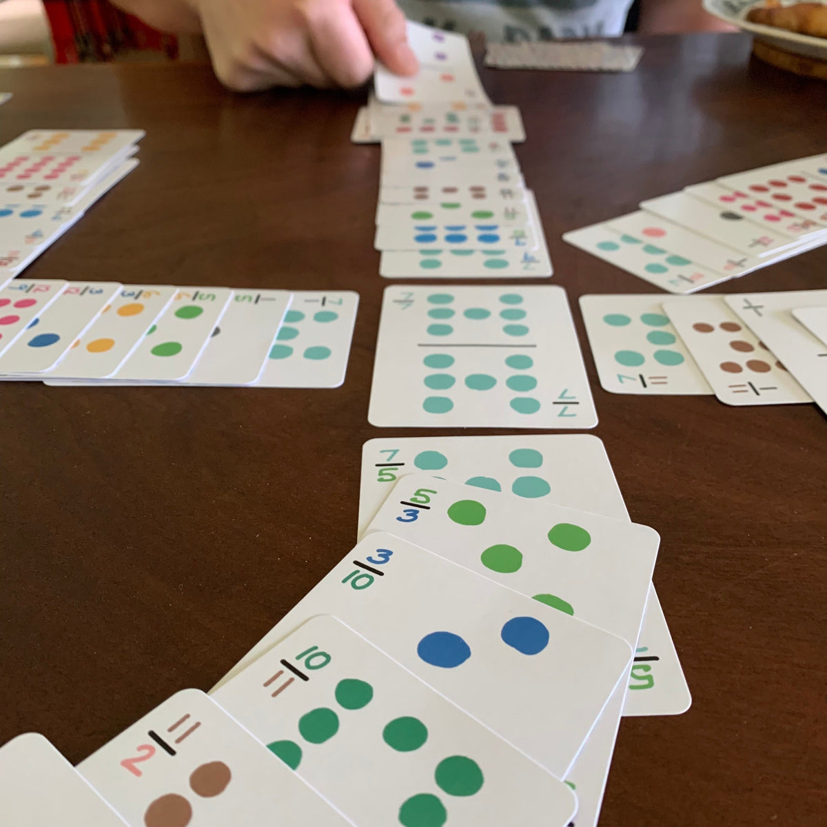 A close-up of a card game in progress on a wooden table, with multiple hands visible arranging and flipping round cards marked with colored dots and numbers.